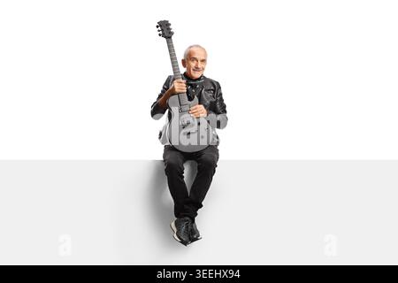 Smiling elderly man in a leather jacket sitting on a panel and holding an electric guitar isolated on white background Stock Photo