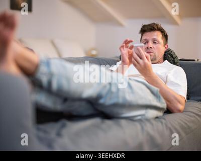 Casual man lying on a gray sofa and using a smartphone at home Stock Photo