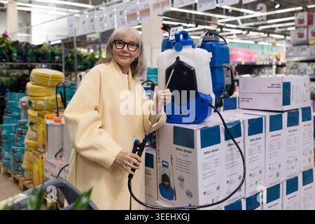Senior woman in glasses and yellow coat holding a battery sprayer in a hardware store Stock Photo