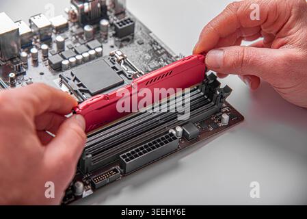 A person's hands installing a red RAM module into a slot on a computer motherboard. Stock Photo