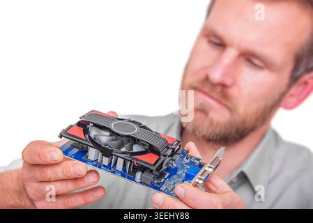 A focused man in a grey shirt holds and inspects a blue computer graphics card with a black cooling fan. Stock Photo