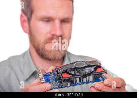A focused male technician in a grey shirt holds and inspects a computer graphics card with a cooling fan. Stock Photo