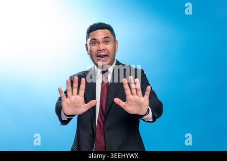 Portrait of an Indonesian professional man in a suit making a stop or denial gesture. Studio shot on blue background representing rejection or warning Stock Photo