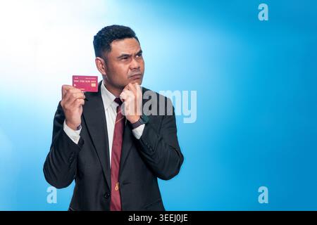 Portrait of an Indonesian businessman in a suit looking up thoughtfully while holding a credit card. Professional studio shot on a blue background. Stock Photo