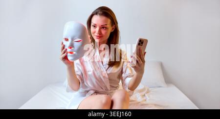 Woman sits on bed with smartphone and LED light therapy mask, soft natural light, white bedding, and minimal background Stock Photo