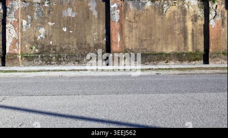Grunge boundary wall with cracked plaster, moss and weeds. Sidewalk and street in front. Urban background for copy space. Stock Photo