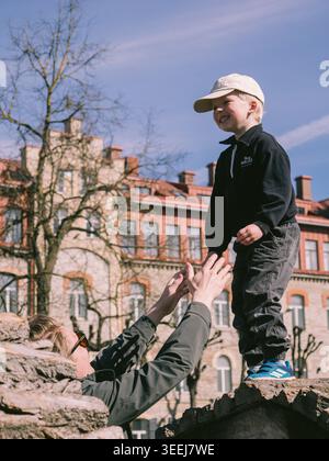 Boy standing on stone wall gives high five to child below in sunny Tallinn Old Town, spring Stock Photo