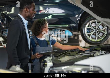 Professional woman mechanic in a modern repair shop using a tablet to explain a car breakdown to a client, an African American businessman. Stock Photo