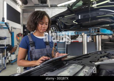 Maintenance of modern vehicles in a successful auto repair shop, a woman mechanic using a digital tablet and manually inspecting a car. Stock Photo