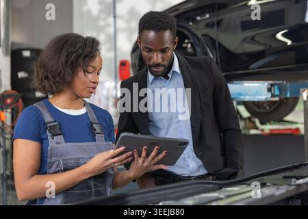 Female mechanic in a professional workshop uses a tablet to review vehicle maintenance checklist together with a client while inspecting the engine. Stock Photo