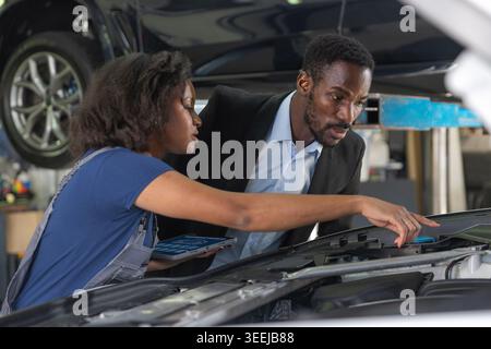 Female mechanic in a professional workshop uses a tablet to review vehicle maintenance checklist together with a client while inspecting the engine. Stock Photo