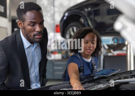 Woman mechanic explaining car problem to client, showing digital diagnostics on tablet. Inclusivity and professionalism in the auto industry. Stock Photo