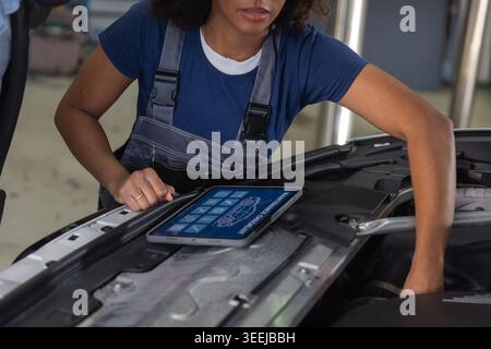 Maintenance of modern vehicles in a successful auto repair shop, a woman mechanic using a digital tablet and manually inspecting a car. Stock Photo