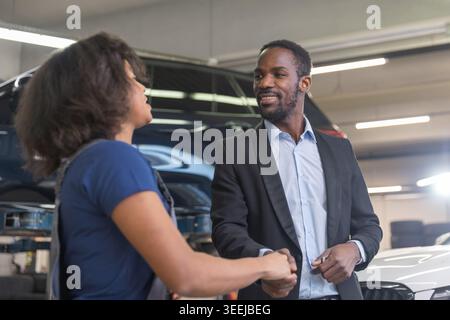 Smiling woman mechanic shaking hands and handing over the car keys to a satisfied, elegant businessman, an auto repair shop client. Stock Photo