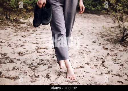 Person walks barefoot on sandy ground while carrying shoes. Earthing and grounding technique, sensory experience outdoors, mental health relief. Stock Photo