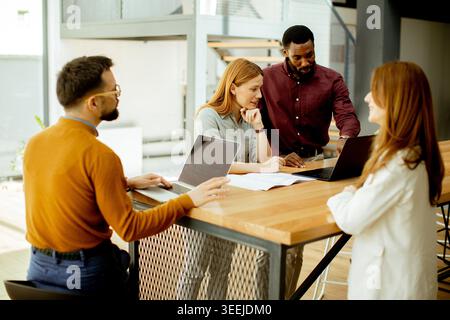 Four people are gathered around a table in a workspace. They discuss a project while looking at laptops. The time appears to be working hours during w Stock Photo