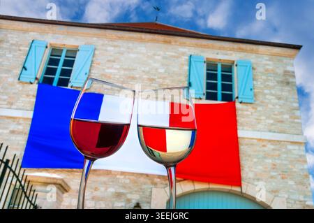 FRENCH WINE TASTING FUN CHEERS Red and White wine glasses lean towards each other against large French flag draped on wall of patriotic French winery Stock Photo