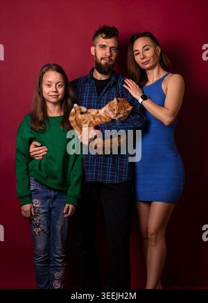 Studio portrait of a modern family—mother, father, daughter—posing with their ginger cat against a burgundy backdrop. Stock Photo