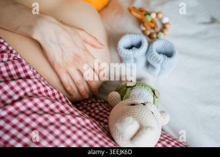 Expecting mother tenderly cradling her baby bump on a bed, surrounded by tiny blue knitted booties, wooden teether and a cuddly crocheted toy in soft light Stock Photo