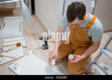 Man kneeling on the floor, carefully reading assembly instructions and attaching parts to a white flat-pack furniture piece, surrounded by tools and components Stock Photo