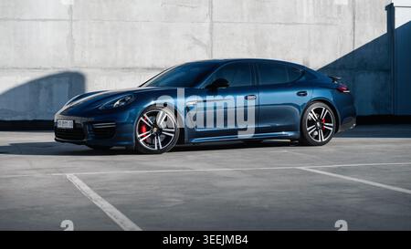blue Porsche Panamera in front of concrete wall on sunny day. Three quarter low-angle front view of german grand tourer in rooftop parking lot. Stock Photo