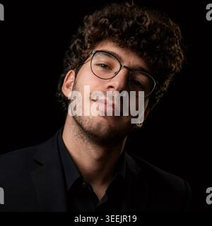 Young man with curly hair, neat beard stubble and glasses in a black suit against a dark studio background, confident slight smile, head and shoulders Stock Photo