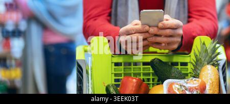 Person Shopping in a Grocery Store Using a Smartphone While Standing Next to a Cart Filled With Food Stock Photo