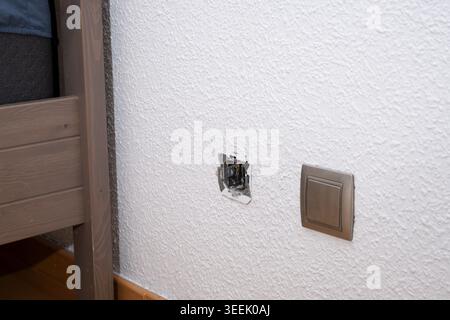 Exposed electrical wiring from a damaged wall socket on a textured white wall Stock Photo