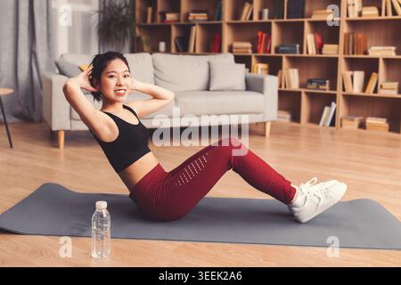 Woman Exercises on Mat in Living Room While Smiling and Doing Sit-Ups in Daylight Stock Photo