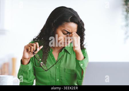 Woman Sitting at Desk With Glasses Holding Her Forehead Due to Stress While Working on Laptop Stock Photo
