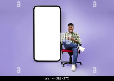 Young Man Sitting in a Chair Using a Smartphone Next to a Large Blank Screen Stock Photo