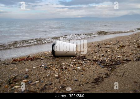 A discarded white paper coffee cup with a black lid lying as litter on a sandy sea beach close-up. Environmental pollution. Stock Photo