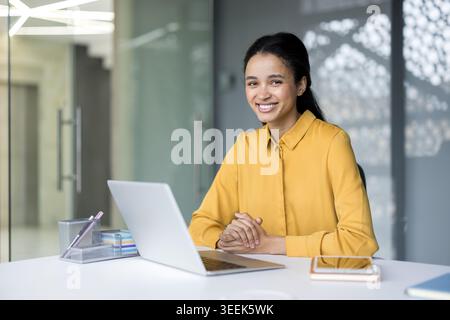 Young female professional beaming at the camera while sitting at her desk with a laptop, reflecting business success, confidence, and a positive corporate work environment Stock Photo