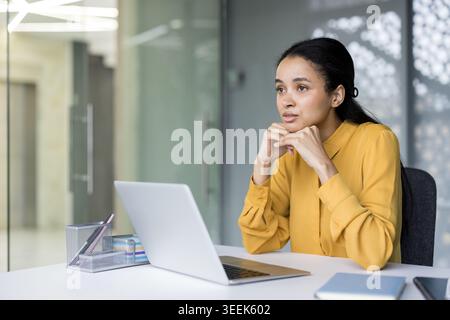 Professional woman in a yellow blouse seated at a modern office desk with laptop, deep in thought as she plans strategy, analyzes ideas and contemplates business solutions Stock Photo