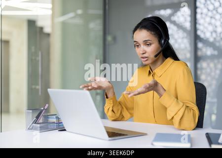 Young african american woman in a yellow shirt wearing a headset, focused on laptop while providing virtual customer support, consulting and problem-solving during a video call Stock Photo