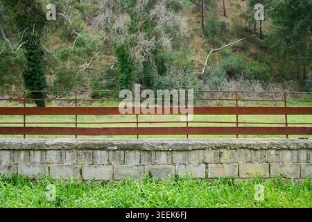 A long rusty metal railing on top of a concrete brick wall in front of a green forest hill background, rural scenery. Stock Photo