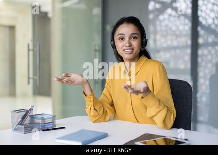 Young mixed race businesswoman wearing a headset and talking during a video call, gesturing with her hands while working from a modern office workspace Stock Photo