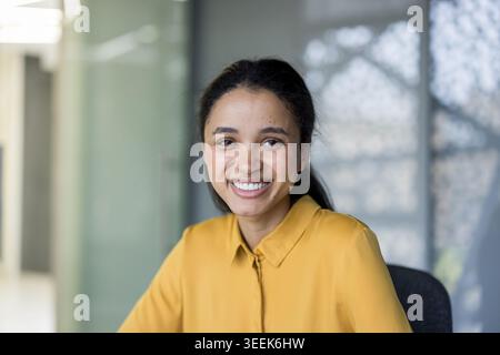 Young mixed race woman smiling at camera, representing happiness, confidence, and professionalism in her thriving career, sitting in a modern corporate office setting Stock Photo