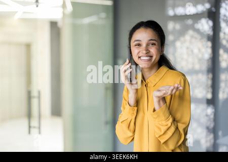 Smiling black woman engaging in a cheerful conversation on her smartphone, discussing business or personal matters while standing in a modern office environment Stock Photo