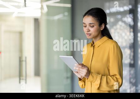 Professional woman using a digital tablet for business tasks while standing in a modern office hallway, focusing on technology, communication, and connectivity Stock Photo