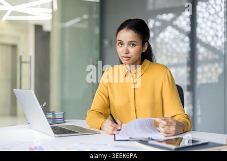 Young professional businesswoman at a modern desk, focused on documents and laptop, confidently reviewing reports and contracts in a contemporary office environment, poised and productive Stock Photo