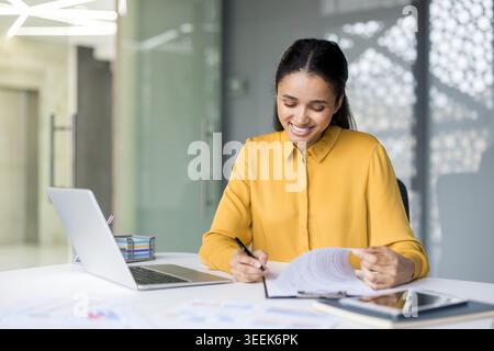Young mixed race businesswoman smiling while reviewing and signing important contractual documents, managing paperwork and responsibilities at a desk in a contemporary office setting Stock Photo