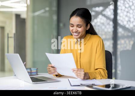Young businesswoman with a cheerful expression reading important documents while sitting at a desk with a laptop, managing her workday in a bright corporate environment Stock Photo