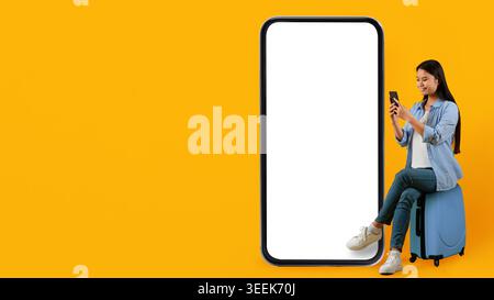 Woman Using Smartphone While Sitting on Suitcase Next to Large Blank Screen Stock Photo