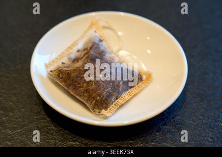 A used herbal tea bag sitting on a small white ceramic saucer over a dark textured slate background, morning routine scene. Stock Photo