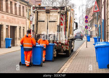 Sanitation workers in orange uniforms collecting blue wheelie bins into a garbage truck on a city street in Germany. Stock Photo