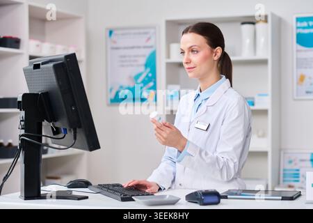 Young female pharmacist checking medicine container while using computer at pharmacy counter, supporting prescription service, retail healthcare marketing, wide shot Stock Photo