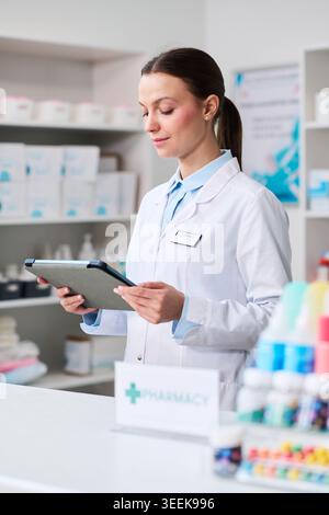 Vertical shot of young pharmacist checking tablet in pharmacy, managing inventory and patient service. Useful for healthcare retail, medication management, digital workflow Stock Photo