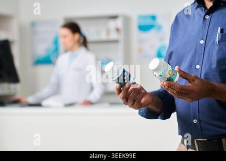Middle aged man comparing supplement bottles in pharmacy while female pharmacist working in background, supporting healthcare retail, product choice, wellness marketing Stock Photo