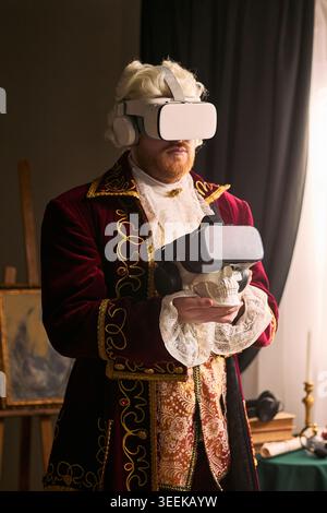 Vertical shot of young man wearing historical costume and using virtual reality headset while holding skull with headset, illustrating innovation meeting heritage for tech campaigns Stock Photo